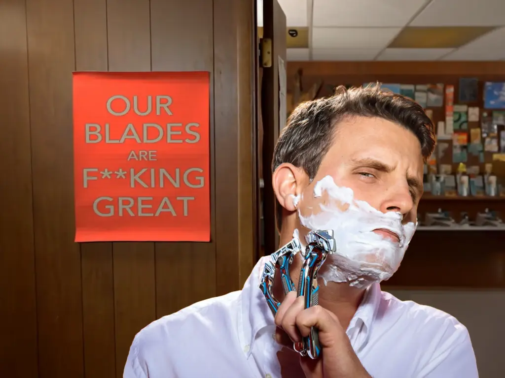 The CEO of Dollar Shave Club, a man, shaves his face with a razor covered in shaving cream in a room with wooden paneling. He holds a modern razor with multiple blades. Behind him, a red sign with white text reads, "OUR BLADES ARE F***ING GREAT." The background includes shelves with various colorful items and a partially open door.